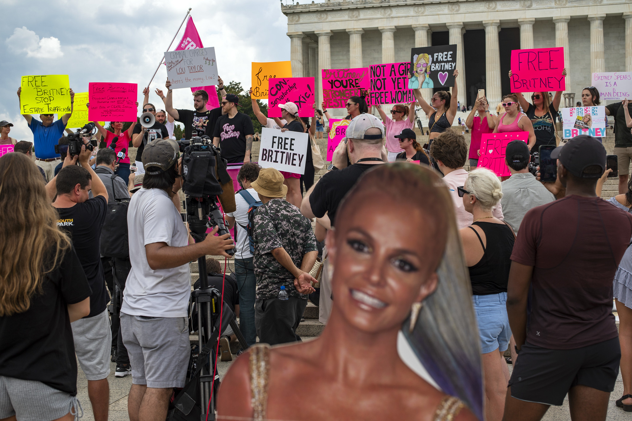 A crowd of Britney Spears supporters hold up signs for the media in front of the Lincoln Memorial. A cardboard Britney is in the foreground.